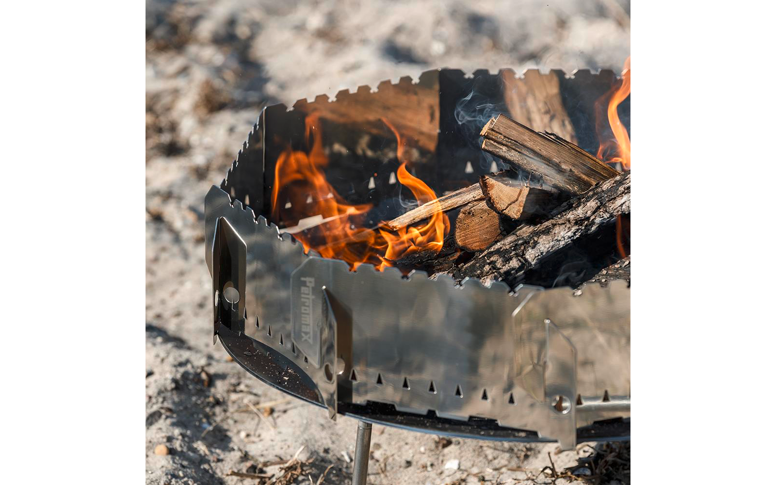 Metallfeuerschale auf kurzen Beinen mit brennenden Holzscheiten und Flammen auf sandigem Untergrund