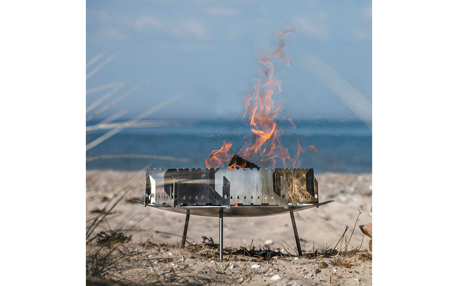 Metall-Feuerschale mit Flammen auf sandigem Strand, Meer im Hintergrund