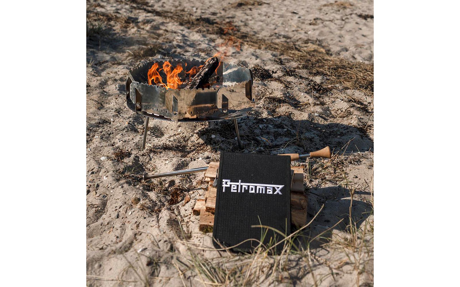Metallbehälter auf Beinen mit brennenden Holzscheiten auf sandigem Untergrund; davor gestapelte Holzscheite, schwarzes Etui mit weißem Schriftzug "Petromax" und eine Zange mit Holzgriff