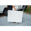 Personne portant une table pliante blanche repliée devant un camping-car sur un tapis extérieur gris à motif