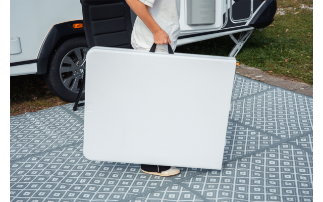 Personne portant une table pliante blanche repliée devant un camping-car sur un tapis extérieur gris à motif