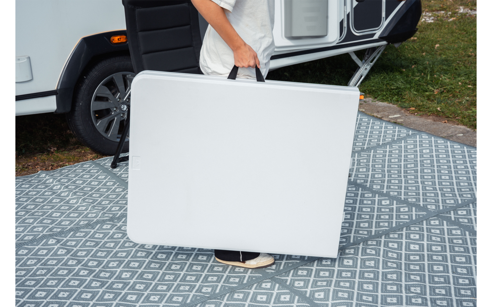 Personne portant une table pliante blanche repliée devant un camping-car sur un tapis extérieur gris à motif