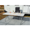 Table pliante blanche sur tapis extérieur à motifs devant une caravane, avec plante en pot, verre, tasse, bougie et carafe en verre dessus; fauteuil de camping noir derrière