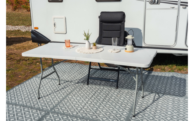 Table pliante blanche sur tapis extérieur à motifs devant une caravane, avec plante en pot, verre, tasse, bougie et carafe en verre dessus; fauteuil de camping noir derrière