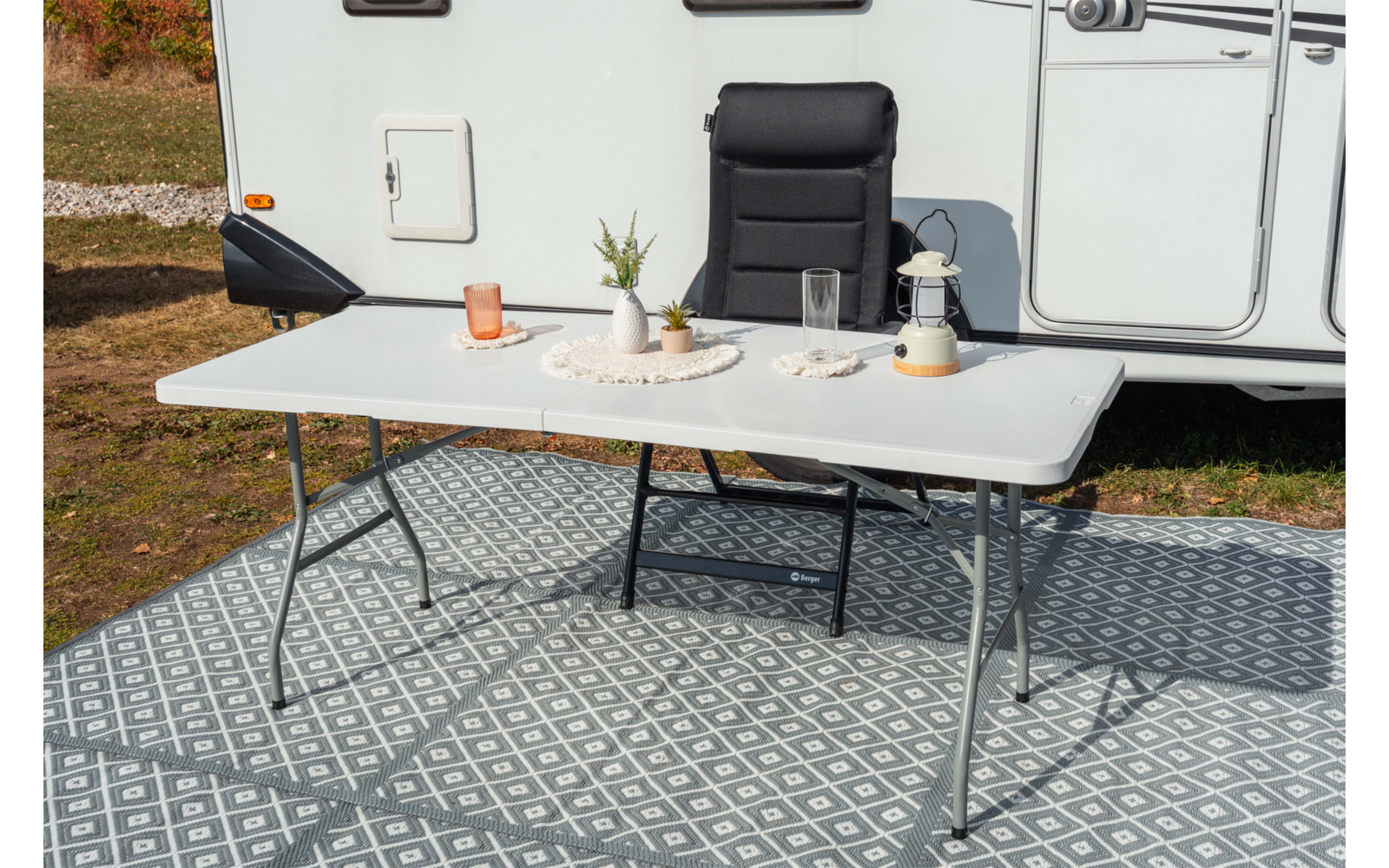 Table pliante blanche sur tapis extérieur à motifs devant une caravane, avec plante en pot, verre, tasse, bougie et carafe en verre dessus; fauteuil de camping noir derrière