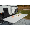 Table pliante blanche avec un verre, coupelle décorative avec plantes et une lanterne, chaise de camping noire, caravane blanche et herbe en arrière-plan