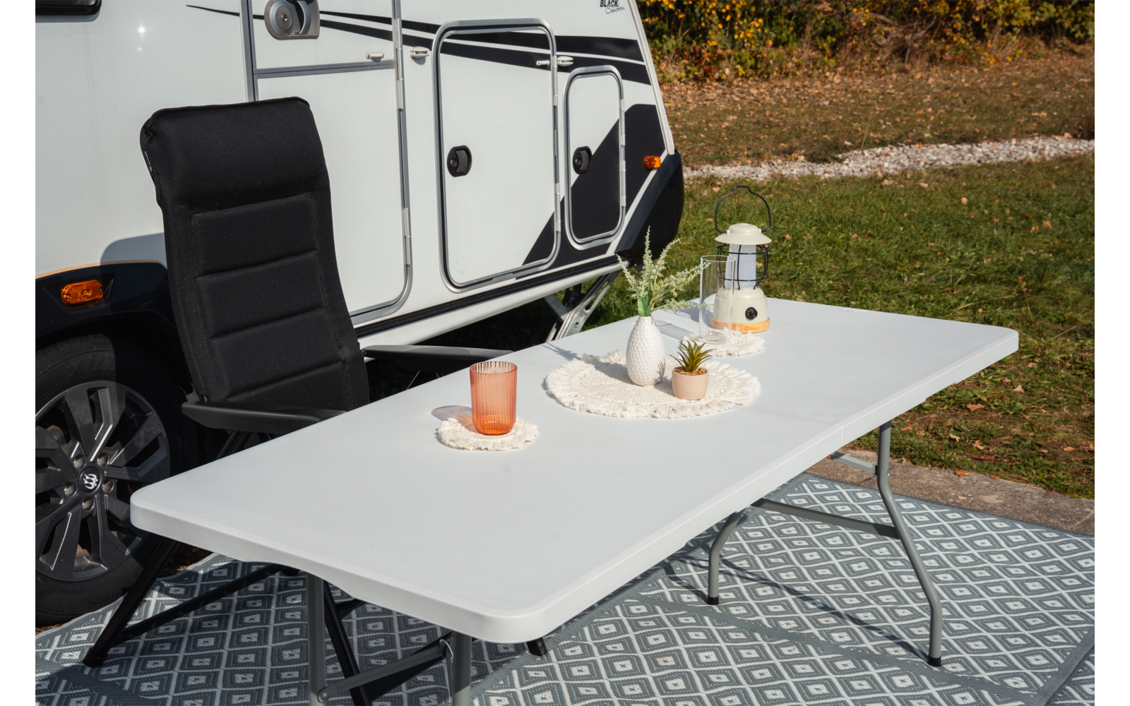 Table pliante blanche avec un verre, coupelle décorative avec plantes et une lanterne, chaise de camping noire, caravane blanche et herbe en arrière-plan