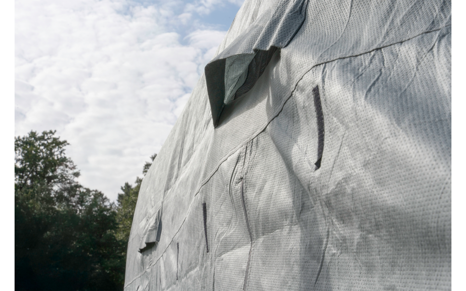 Building facade almost completely covered with a gray tarp; visible seams and folds; cloudy sky and trees at left