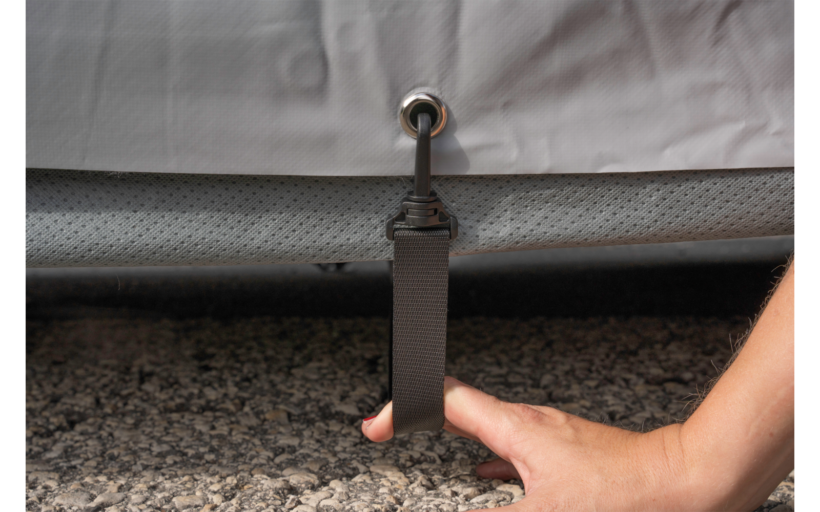 Hand touching a black strap with metal ring attached to the underside of a vehicle, gravel ground below