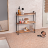Dreistöckiges Holzregal mit schwarzem Metallgestell auf Arbeitsplatte neben Waschbecken; auf den Böden Flaschen, Nagellack, Cremedose, Seifenschale und Glas; Spiegelrahmen links, Jalousie rechts, geöffnetes Puderkompakt vorne