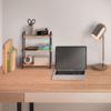 Holztisch mit offenem Laptop (schwarzer Bildschirm), rechts eine Metall-Schreibtischlampe, links ein kleines Regal mit Schere, Stifthalter, Kamera und Klebeband; Bücher und Ordner am linken Rand