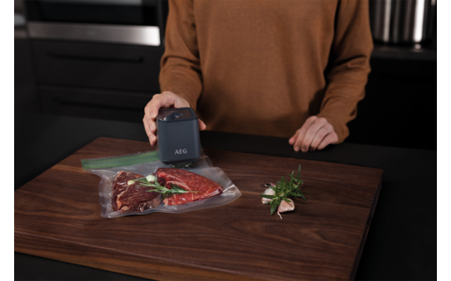 Hände einer Person über einem Holzbrett, ein kleines elektrisches Vakuumiergerät mit Aufschrift "AEG" versiegelt ein rohes Steak in einem Plastikbeutel; daneben Kräuterzweige und Knoblauchzehen, dunkle Küchenarbeitsfläche im Hintergrund
