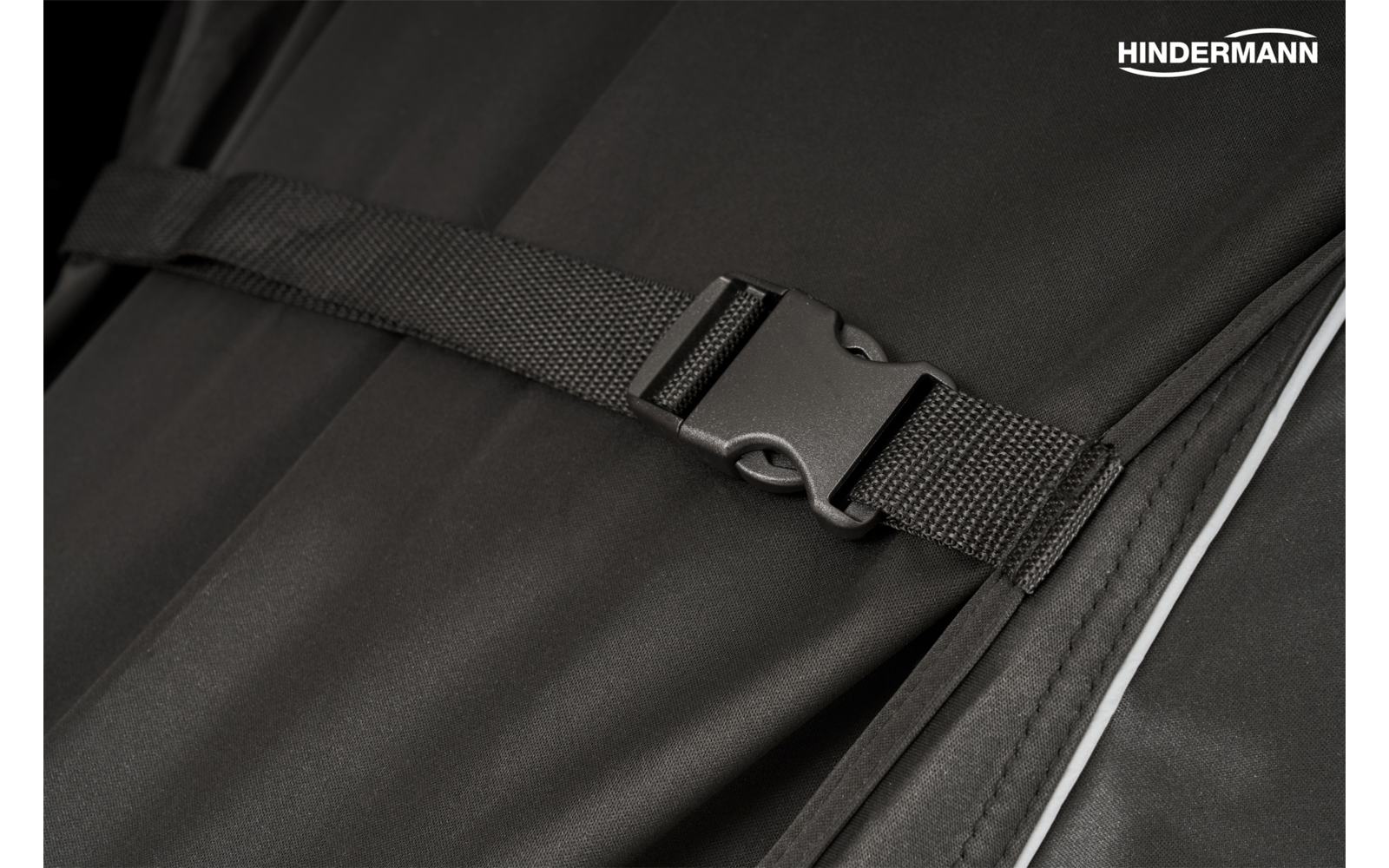 Nahaufnahme einer schwarzen Kunststoff-Steckschnalle an einem schwarzen Gurt auf schwarzem Stoff, weiße Paspel rechts, weißes Markenlogo 'HINDERMANN' oben rechts