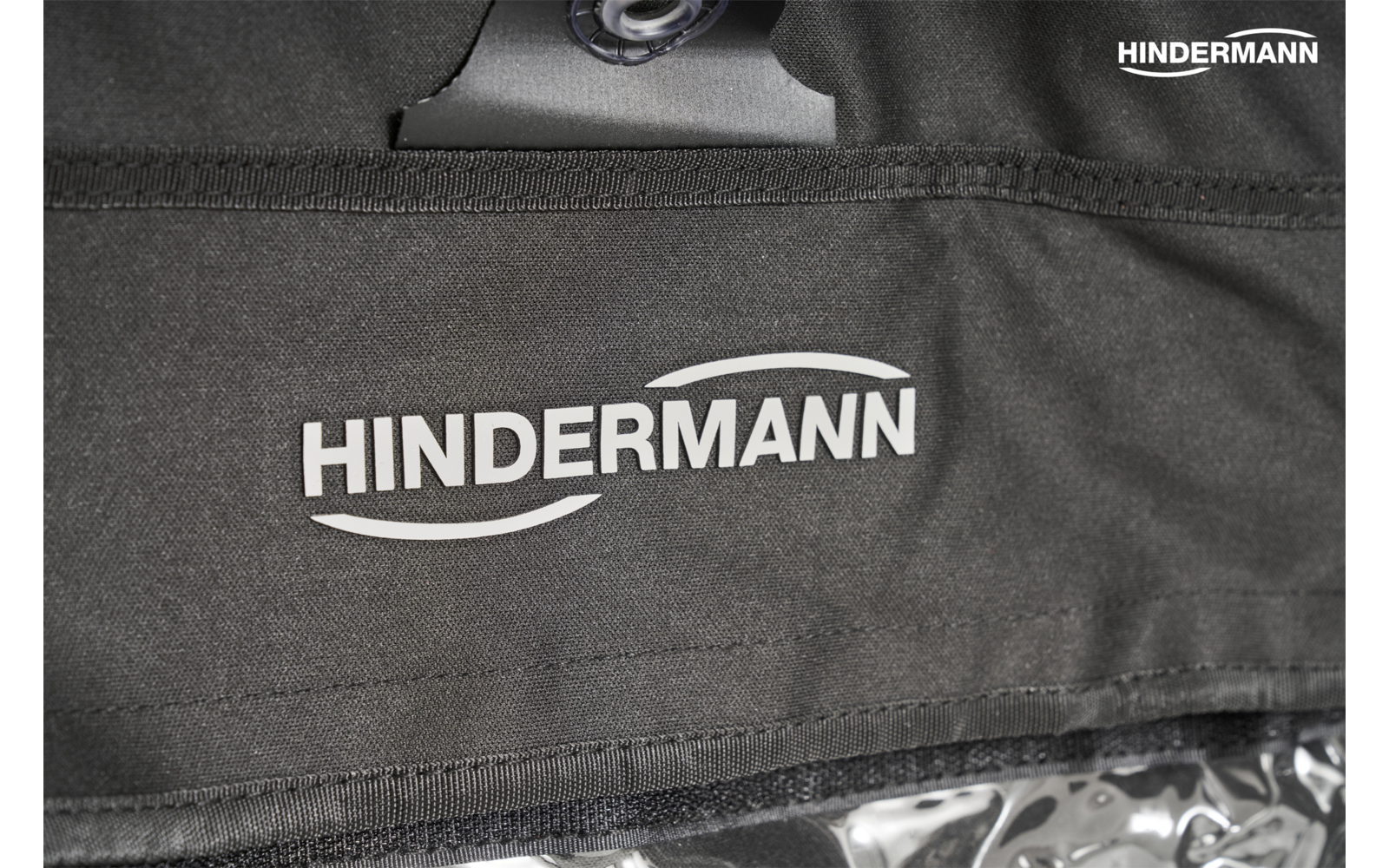 Schwarzer Stoff mit großem und kleinem weißen Schriftzug 'HINDERMANN', sichtbare Naht, Metallknopf oben und transparente Kunststoffkante unten