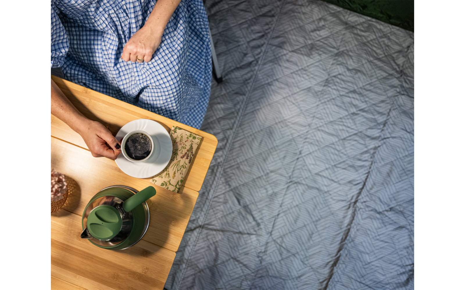 Draufsicht: Bett mit grauer Steppdecke rechts, links ein Teil einer sitzenden Person in blau kariertem Kleid; auf einem Holztablett eine Tasse mit dunklem Getränk auf Untertasse, eine grüne Teekanne, Gebäck und ein Blumenstoff