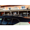 Dach eines hellen Autos mit schwarzem Dach und montierten Querträgern; Spiegelungen in den Fensterscheiben, im Hintergrund ein zweistöckiges Backsteingebäude mit Balkonen und Schaufenstern