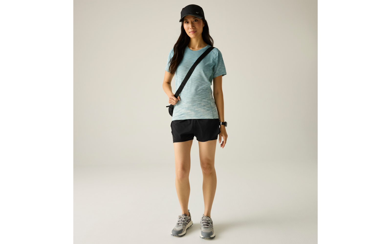 Personne debout sur fond clair, portant un t‑shirt vert menthe, un short noir, une casquette noire, des baskets grises, un sac bandoulière et une montre connectée