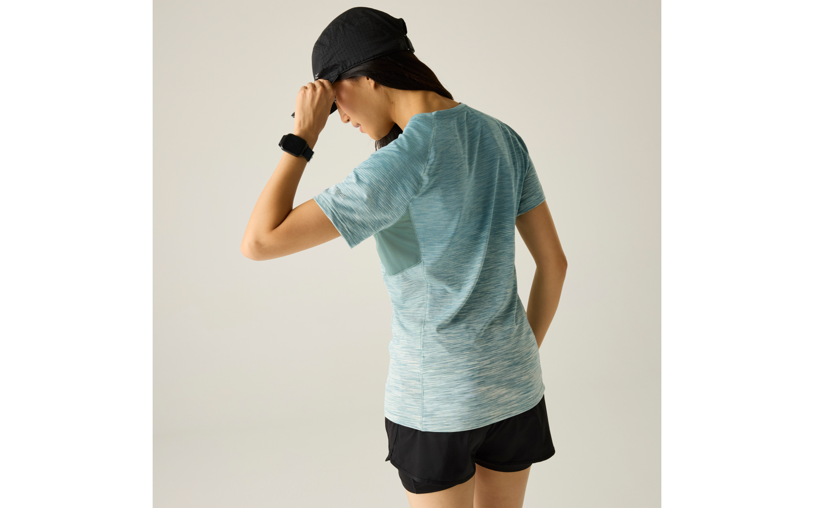 Personne vue de dos, porte un t‑shirt bleu clair à manches courtes, des shorts noirs et une casquette noire; main gauche sur la casquette, montre connectée au poignet gauche; fond uni clair