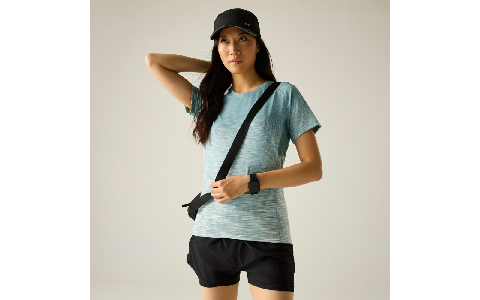 Personne devant un fond gris clair uni, portant une casquette noire, un t‑shirt de sport turquoise à manches courtes, un short noir, une bandoulière noire et une montre connectée ; une main tient la bandoulière, l’autre est derrière la tête, regard vers la droite