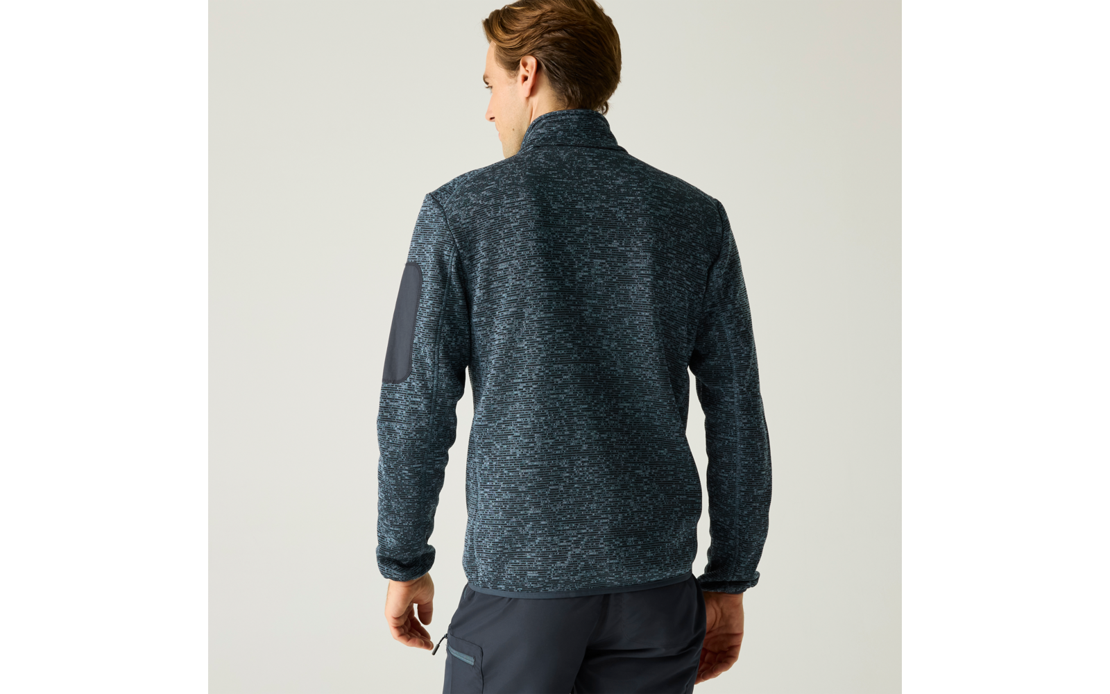 Rückenansicht eines Mannes in dunkelblau melierter Fleecejacke mit hohem Kragen, Ellbogenaufnäher am linken Ärmel und dunkler Hose vor hellem Hintergrund