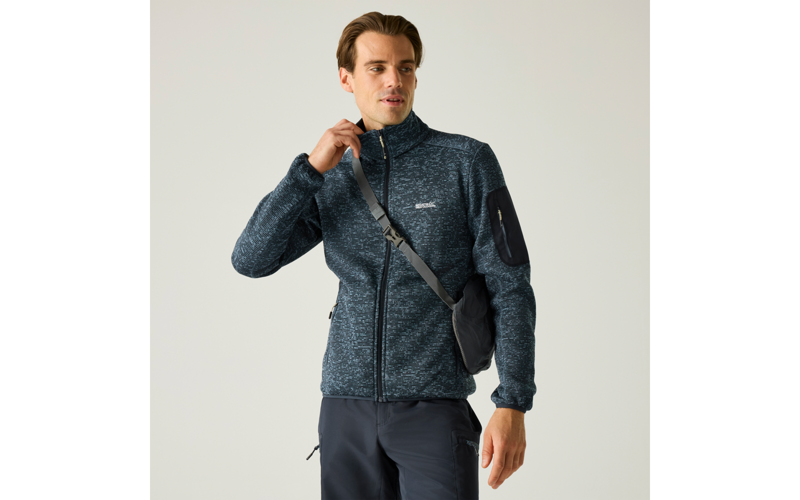 Person mit kurzem Haar trägt dunkelblauen, melierten Zip‑Fleece mit hohem Kragen, eine Umhängetasche und dunkle Hose, steht vor hellem Hintergrund, rechte Hand am Reißverschluss