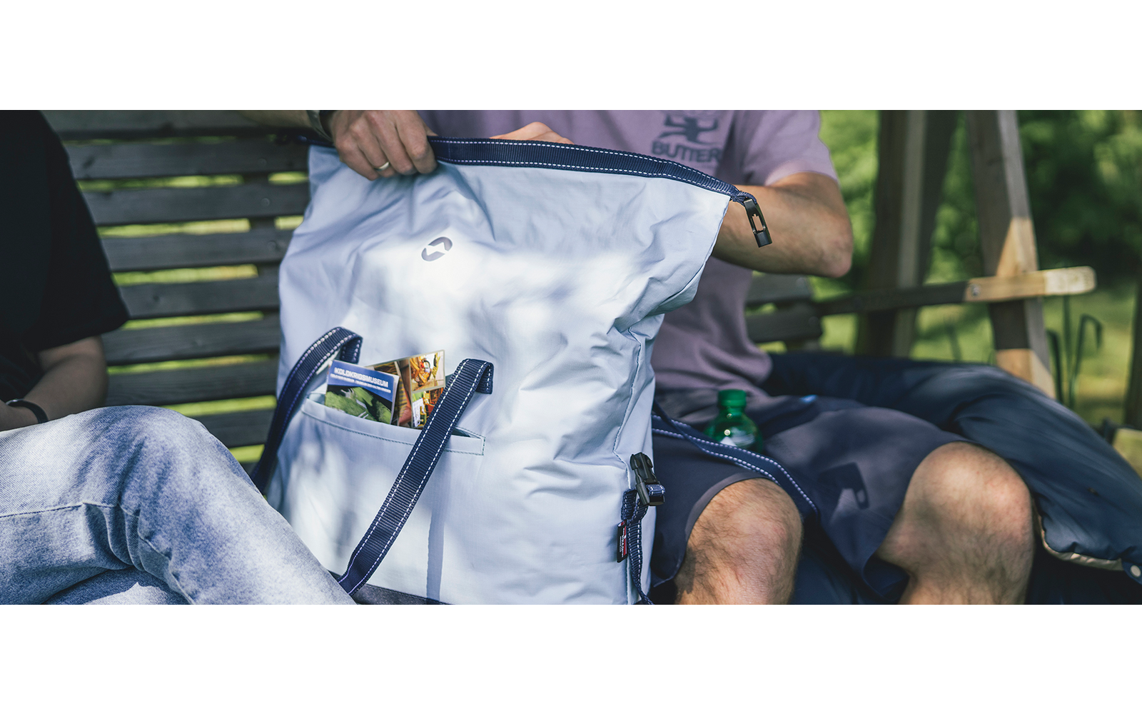 Weißer Rucksack mit offenem Vordertaschenfach, darin sichtbare Snacks; zwei sitzende Personen nur mit Beinen und Hüften sichtbar, eine Hand hält den Rucksack; Holzbank und Wiese im Hintergrund