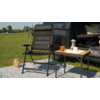 Black folding chair on a gray mat next to a small table with a tray, orange mug and white lantern; open car trunk with pillows and a blanket; grass in the background