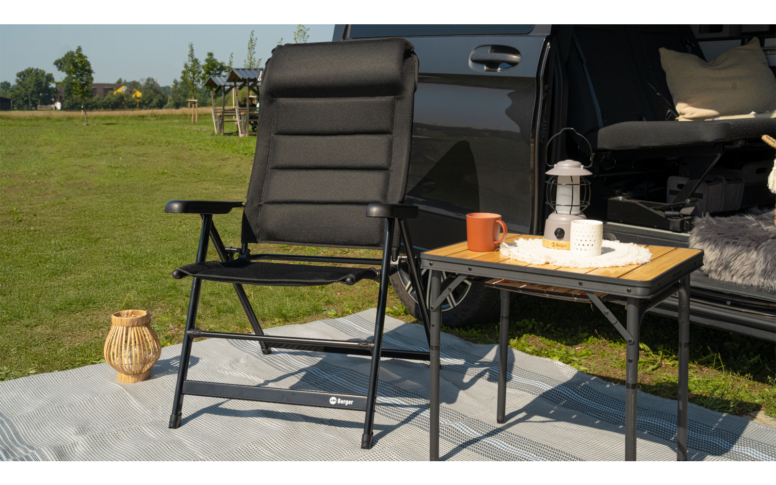 Black folding chair on a gray mat next to a small table with a tray, orange mug and white lantern; open car trunk with pillows and a blanket; grass in the background