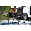 Fauteuil pliant noir rembourré sur tapis à motifs devant une kitchenette de caravane ouverte, à côté d'une petite table avec de la vaisselle, herbe et arbres en arrière-plan