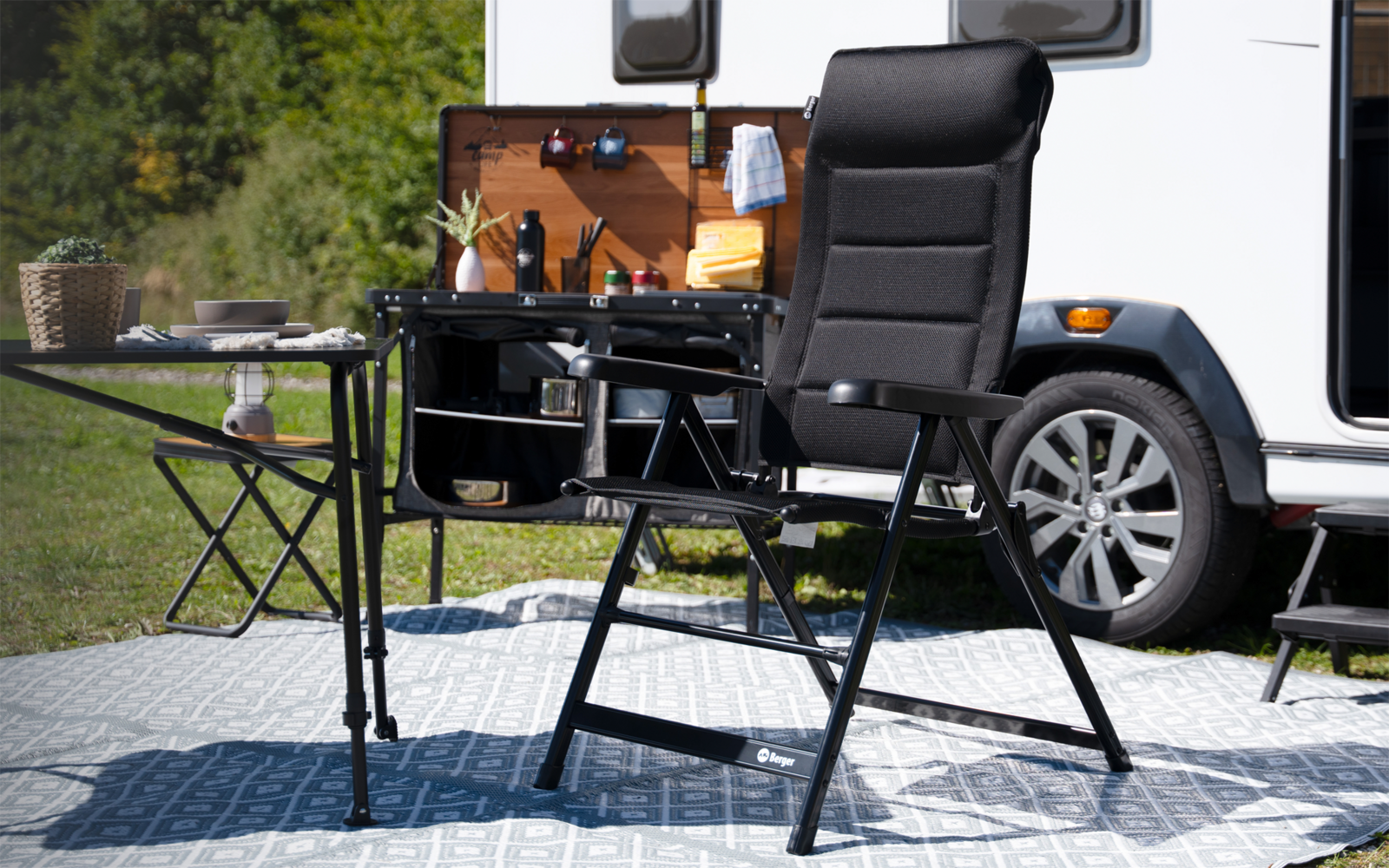 Fauteuil pliant noir rembourré sur tapis à motifs devant une kitchenette de caravane ouverte, à côté d'une petite table avec de la vaisselle, herbe et arbres en arrière-plan
