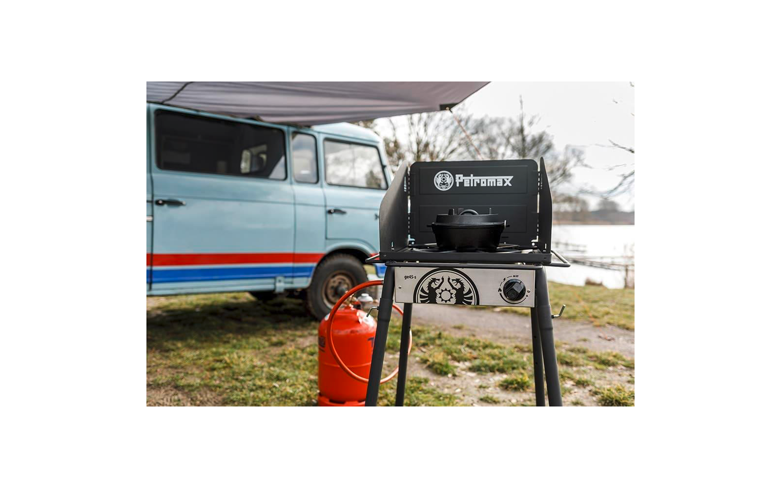 Schwarzer Petromax-Gaskocher auf vier Beinen mit Gusseisentopf, rote Propangasflasche daneben, hellblauer Campingbus mit roter und blauer Streifen im Hintergrund, Planendach darüber, Gras und Bäume im Hintergrund