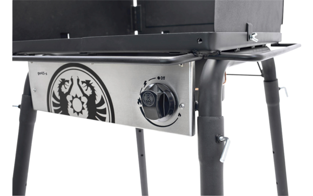 Nahaufnahme eines Grills: Edelstahlfront mit rundem Drehknopf, dekorativem Emblem, schwarzem Gestell und zwei sichtbaren Beinen