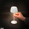 Rechte Hand mit Ring hält den Schaft einer kleinen weißen Tischlampe mit geriffeltem Lampenschirm; Lampe leuchtet vor dunklem Hintergrund. Unten links Schriftzug 'AI GENERATED'