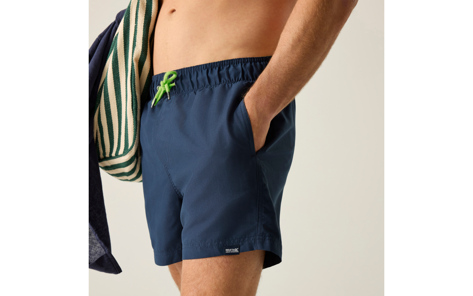 Oberkörper einer Person ohne Oberbekleidung, trägt dunkelblaue Badeshorts mit grünem Kordelzug, rechte Hand in Tasche, gestreiftes Handtuch über der Schulter