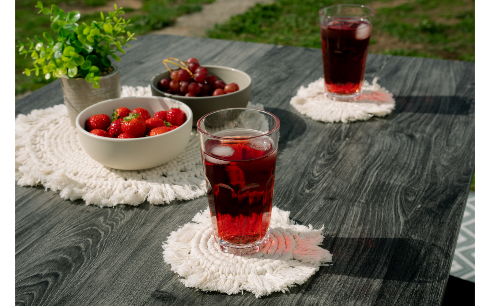 Mesa de madera oscura con dos vasos de bebida roja con hielo sobre posavasos blancos, dos cuencos con fresas y cerezas y una pequeña planta verde en maceta; césped al fondo
