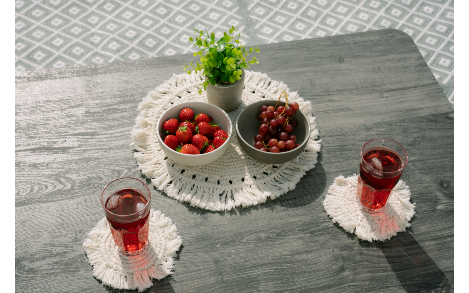 Mesa de madera gris con un tapete blanco de ganchillo, dos cuencos pequeños grises con fresas y uvas rojas, una pequeña planta en maceta verde y dos vasos con líquido rojo sobre posavasos blancos