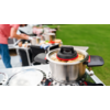 Casserole avec couvercle ouvert sur un réchaud extérieur, une personne et du matériel de camping sont visibles en arrière-plan