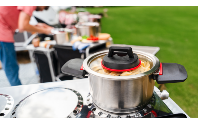 Casserole avec couvercle ouvert sur un réchaud extérieur, une personne et du matériel de camping sont visibles en arrière-plan