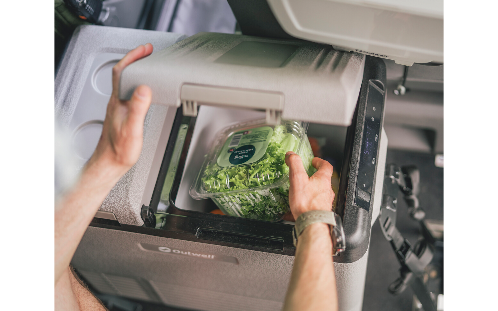 Zwei Hände schieben eine durchsichtige Plastikbox mit grünen Salatblättern in ein geöffnetes Fach im Armaturenbrett eines Autos; rundes Etikett auf dem Deckel; Armbanduhr am Handgelenk