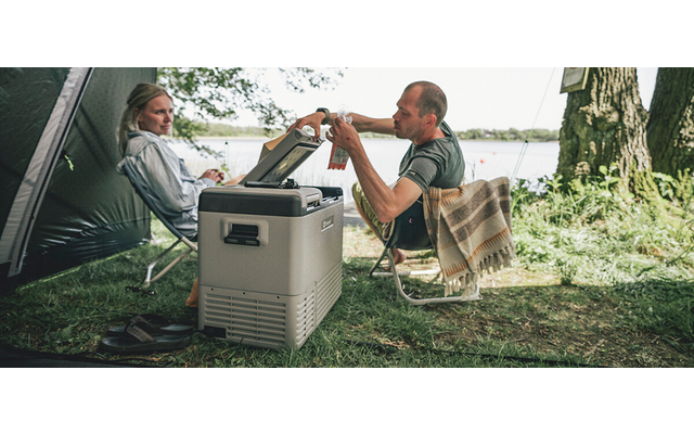 Zwei Personen sitzen auf Campingstühlen neben einem Zelt und einer tragbaren Kühlbox an einem See; eine Person reicht der anderen eine Flasche; Baum, Gras und Wasser im Hintergrund