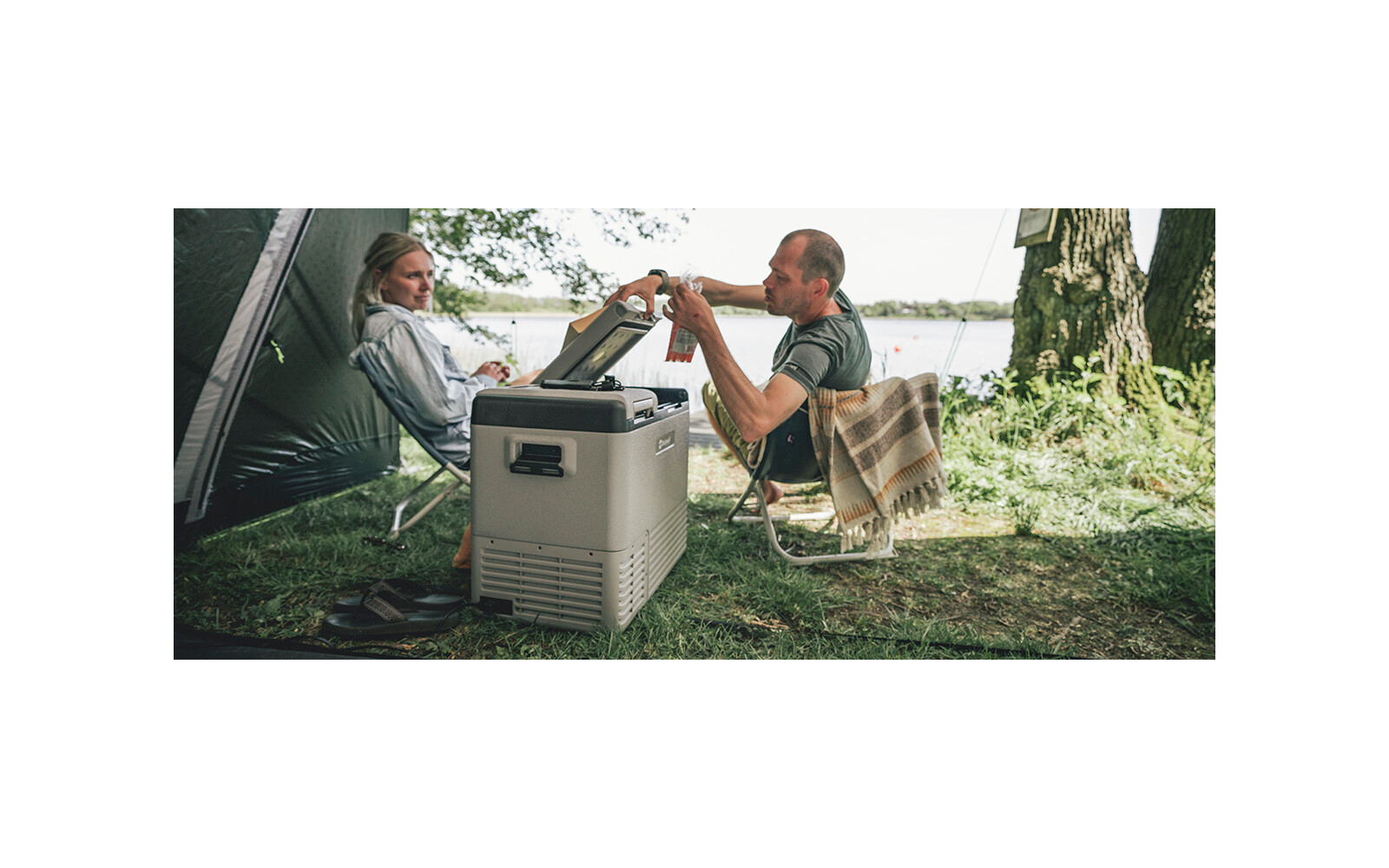 Zwei Personen sitzen auf Campingstühlen neben einem Zelt und einer tragbaren Kühlbox an einem See; eine Person reicht der anderen eine Flasche; Baum, Gras und Wasser im Hintergrund