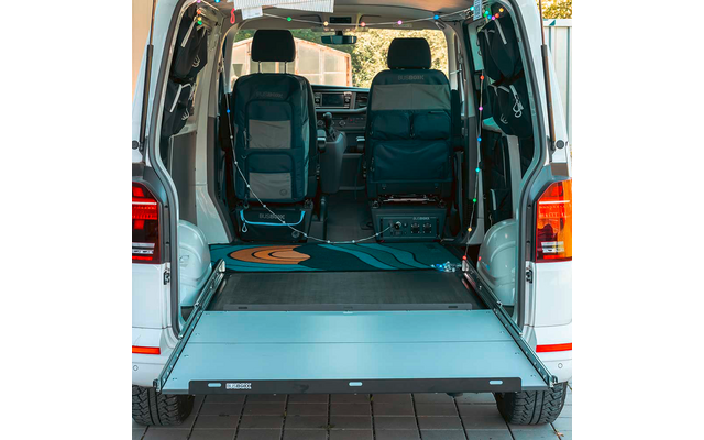 Open rear of a white van showing two front seats, extended loading platform/ramp, blue-and-orange patterned rug, string lights, electronic box with cables on the floor