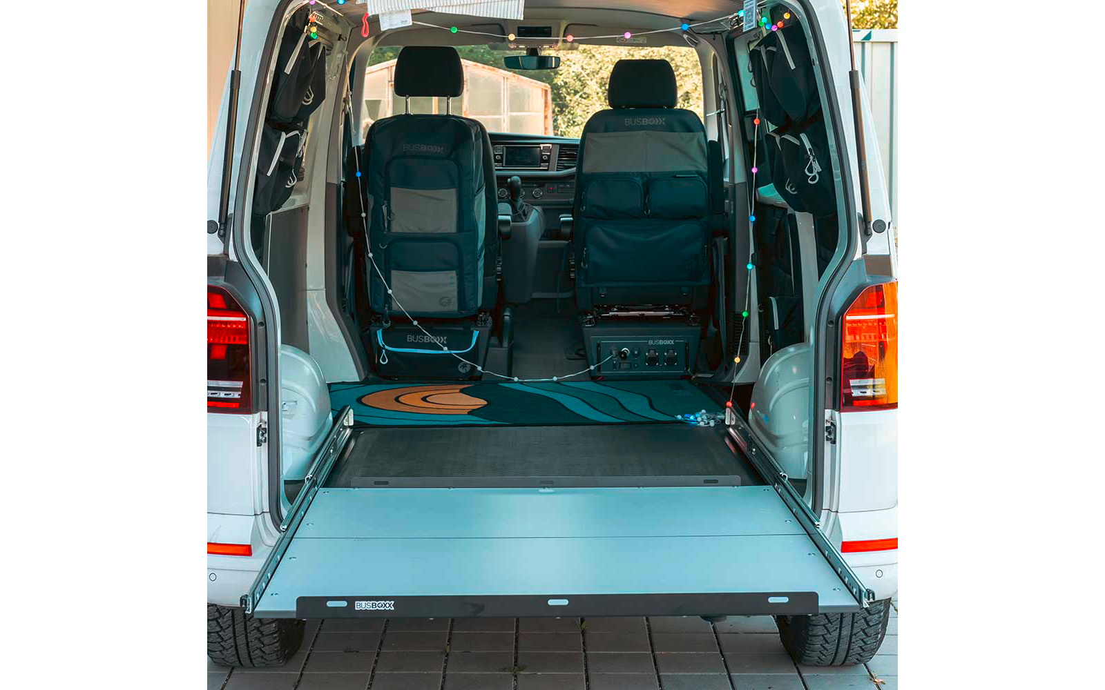 Open rear of a white van showing two front seats, extended loading platform/ramp, blue-and-orange patterned rug, string lights, electronic box with cables on the floor