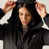 Close-up of a woman lifting the hood of a black jacket with both hands, looking down; a light pink zip-up top and a white logo on the left chest are visible