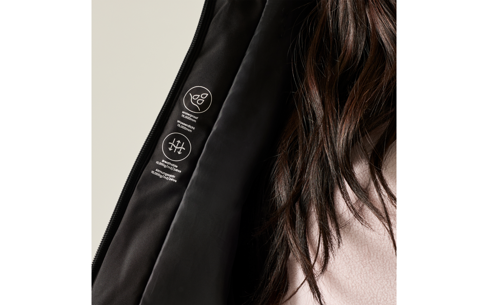 Close-up of a black jacket lining with two white pictograms and small printed text, visible zipper edge, long dark hair on the right and a pink sweater, light background
