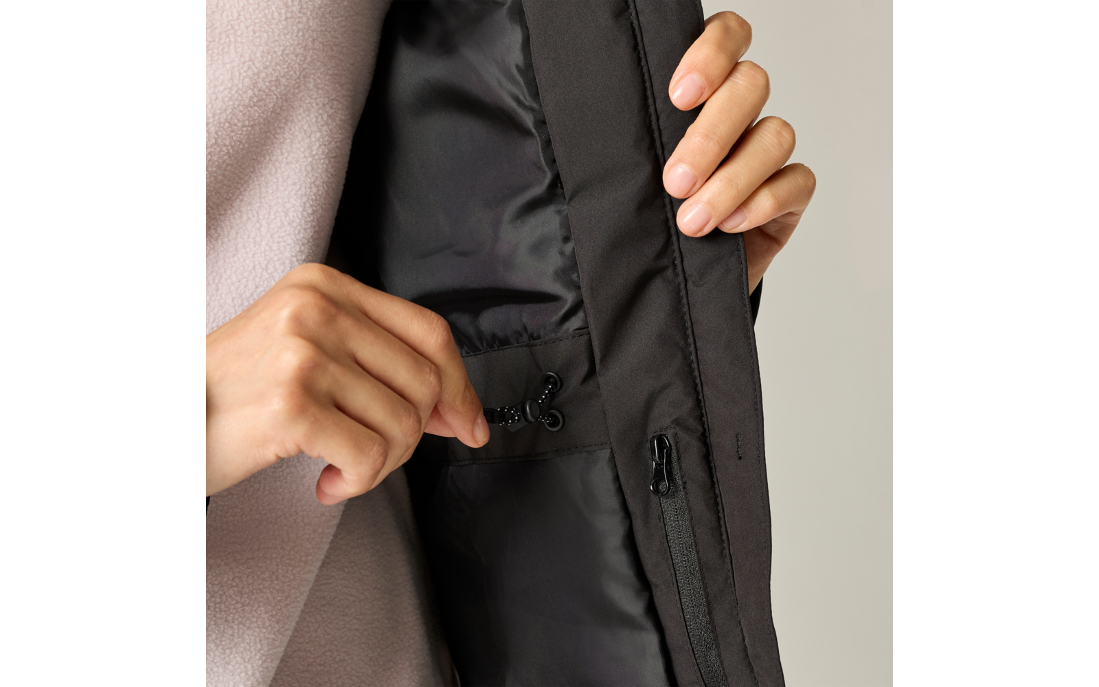 Two hands holding a black drawcord with a cord lock inside a black jacket; visible zipper placket and beige garment underneath