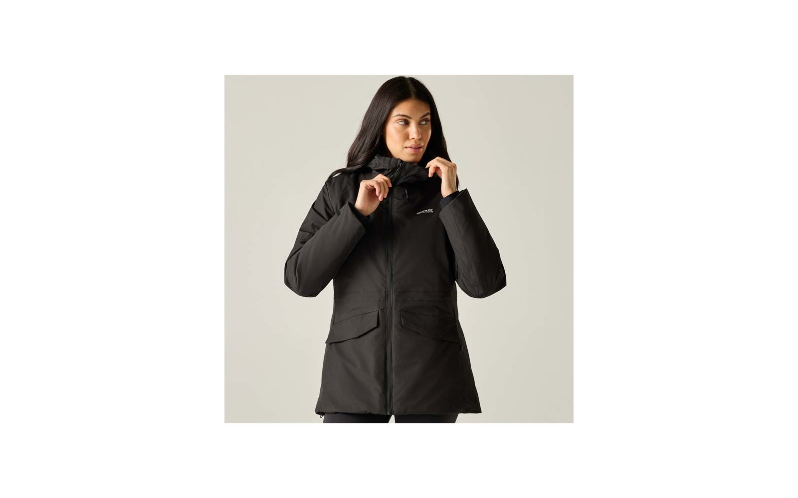 Person with long dark hair wearing a black parka, holding the collar with both hands; two front flap pockets, small chest logo, light background
