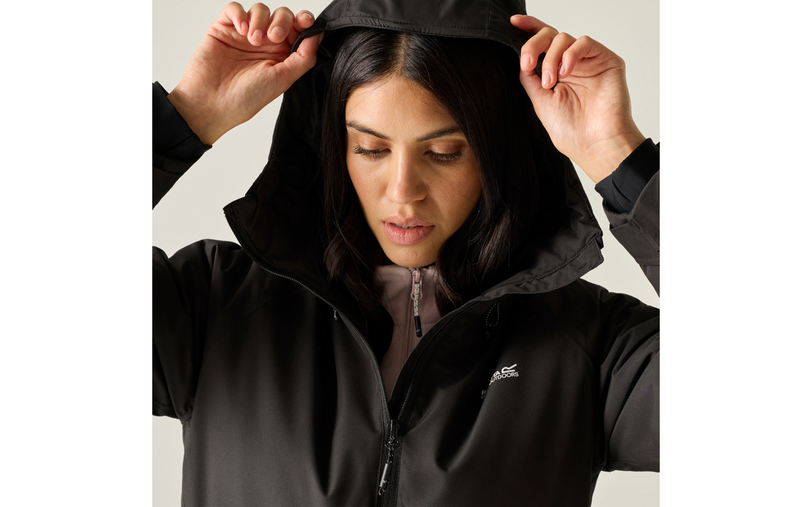 Close-up of a woman lifting the hood of a black jacket with both hands, looking down; a light pink zip-up top and a white logo on the left chest are visible