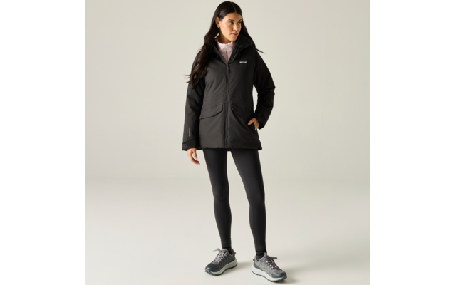 Person stands against a light background wearing a black hooded jacket, black leggings and gray sneakers, light shirt collar visible, long dark hair, one hand in the jacket pocket, looking to the right