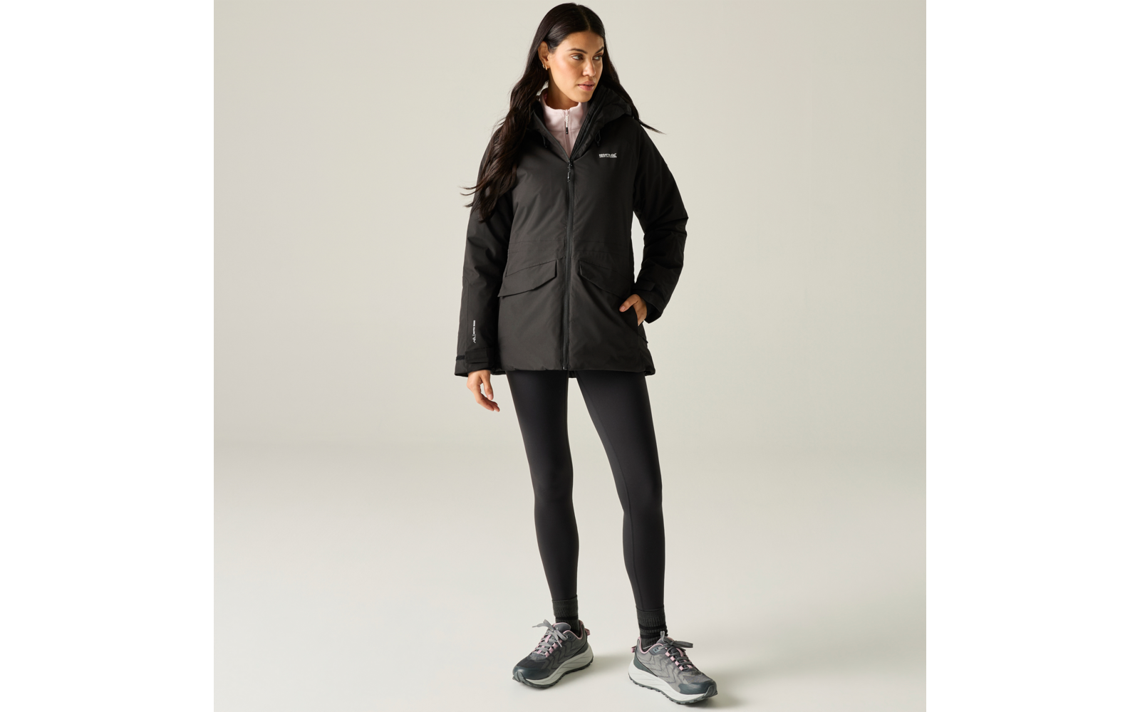 Person stands against a light background wearing a black hooded jacket, black leggings and gray sneakers, light shirt collar visible, long dark hair, one hand in the jacket pocket, looking to the right